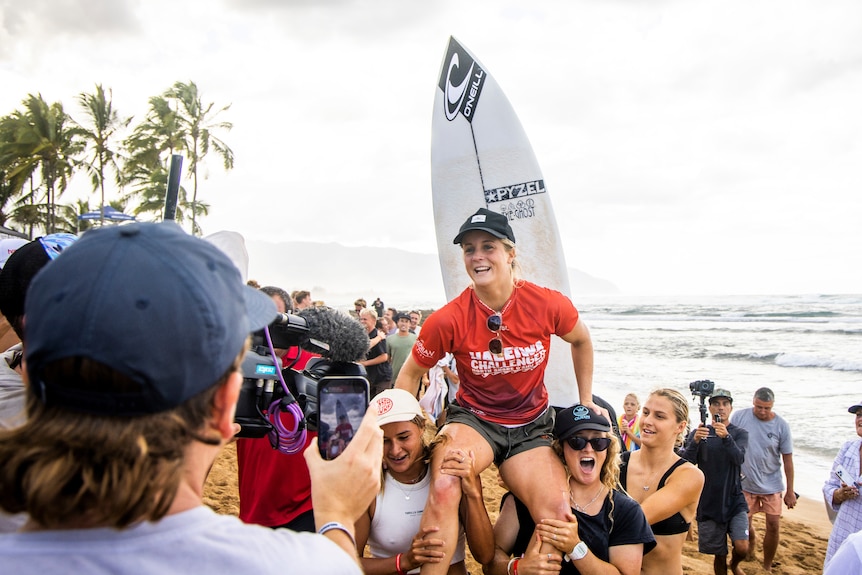 Woman in red smiling, being carried on shoulders, with surfboard behind her