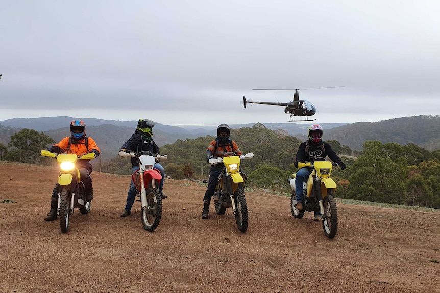 Four motorbikes and their riders on top of a hill with a helicopter flying above.