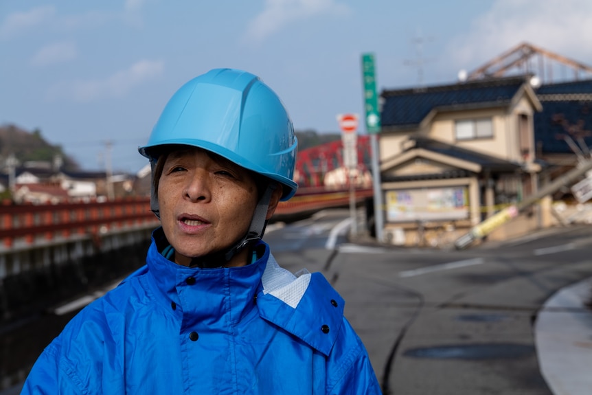 A woman is pictured wearing a blue raincoat and a blue construction helmet. Behind her is damage from the earthquake.