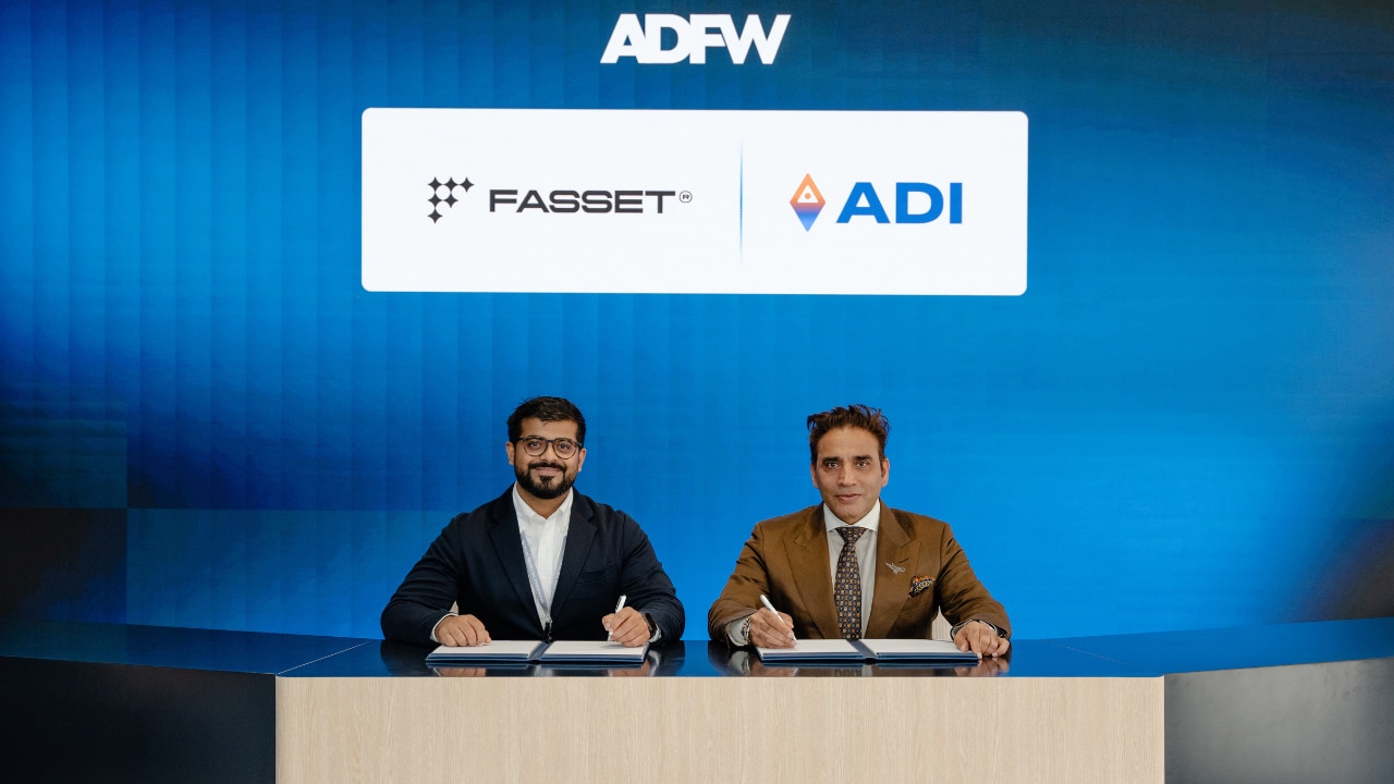 Two men, sitting beneath ADI and Fasset logos, sign documents on a desk.