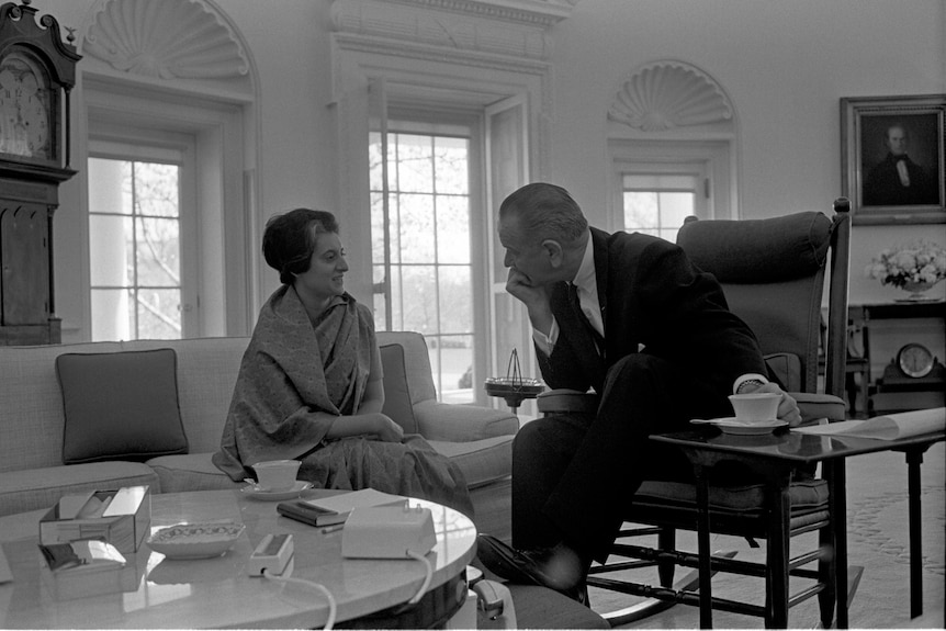 Lyndon B Johnson in a suit sits in a chair leaning his chin on his hand, talking to Indira Gandhi on a couch