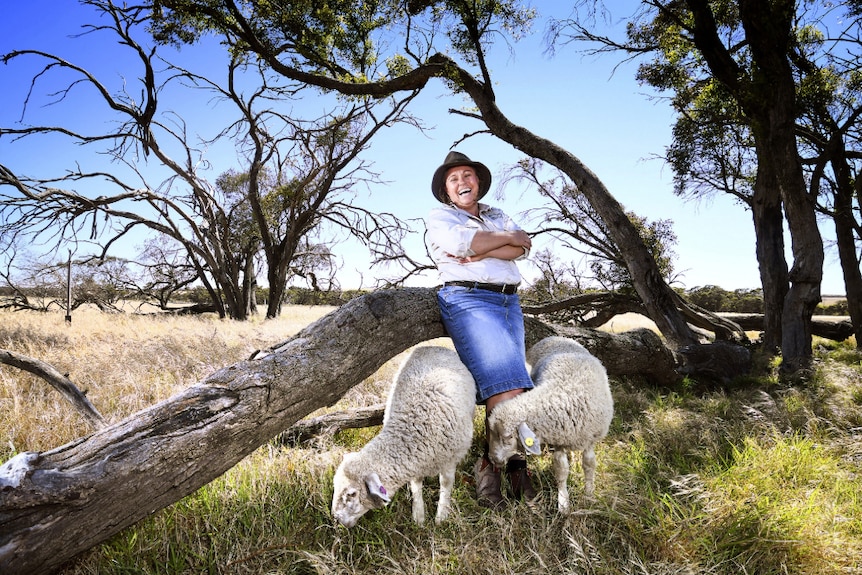 Suzannah Moss-Wright stands beside a tree, with two lambs grazing around her legs.