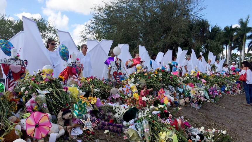 Seventeen people dressed as angels stand near a memorial of flowers.