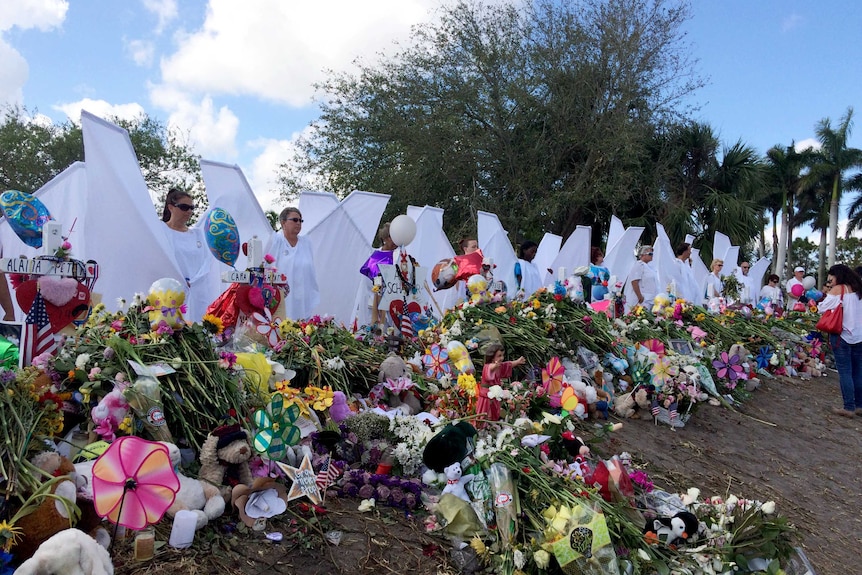 Seventeen people dressed as angels stand near a memorial of flowers.