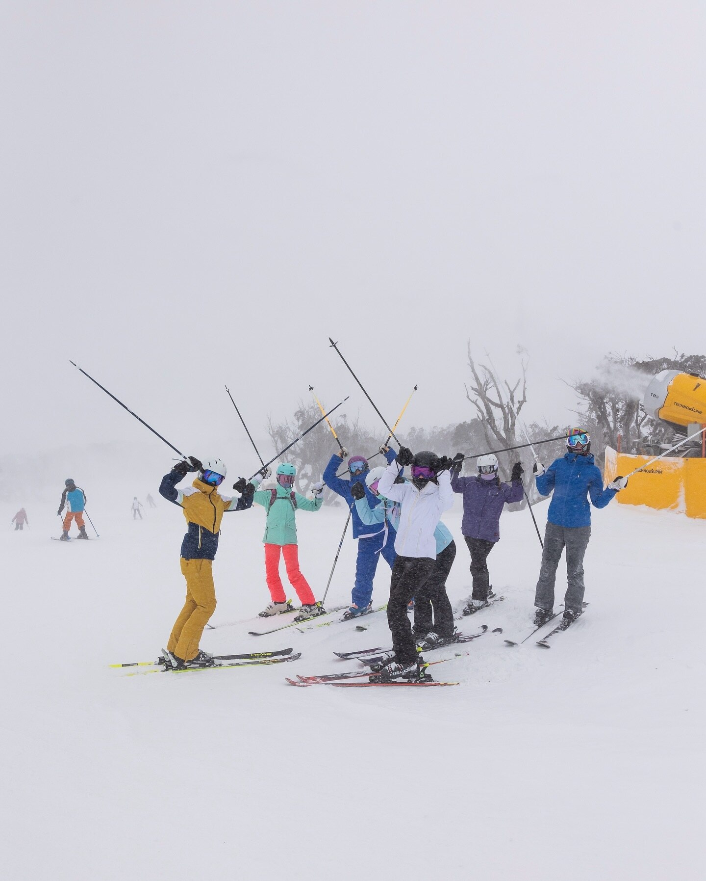 A group of six skiers stand with their poles in the air in the snow field wearing colourful ski suits