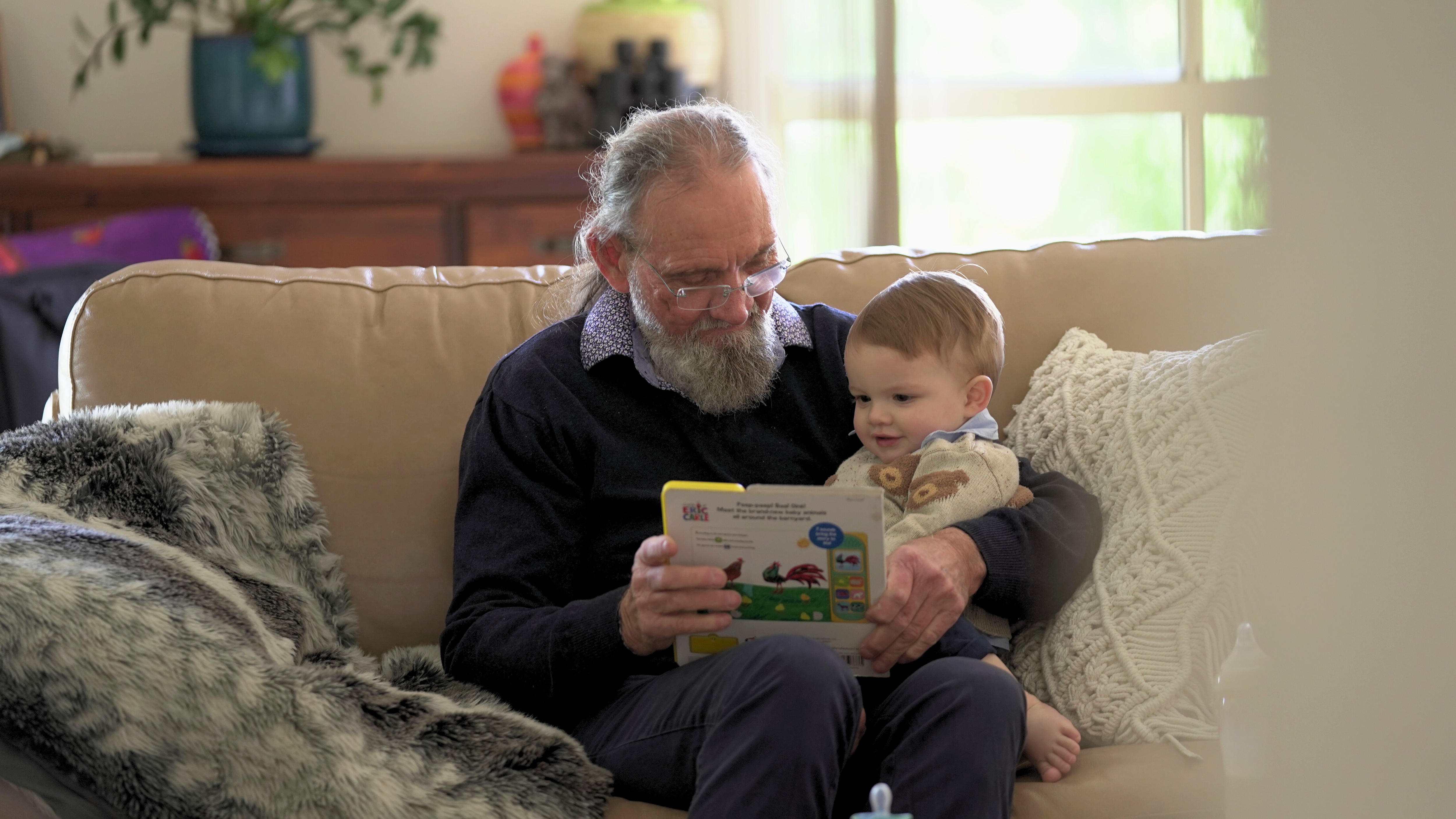 Gregory, a man with a grey beard and glasses reads to his toddler son who sits on his lap.