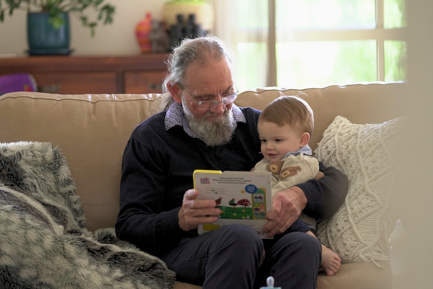 Gregory, a man with a grey beard and glasses reads to his toddler son who sits on his lap.