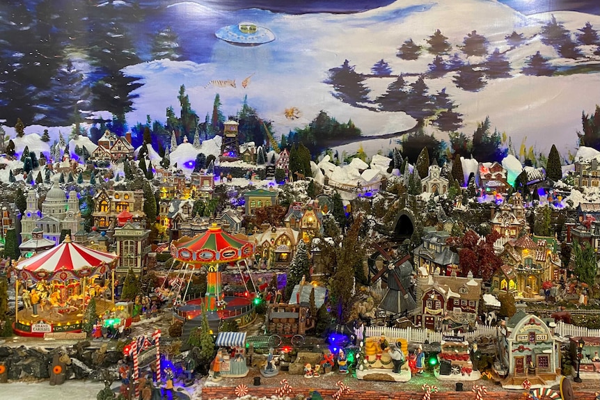Miniature Christmas village with back drop of snow and pine trees.
