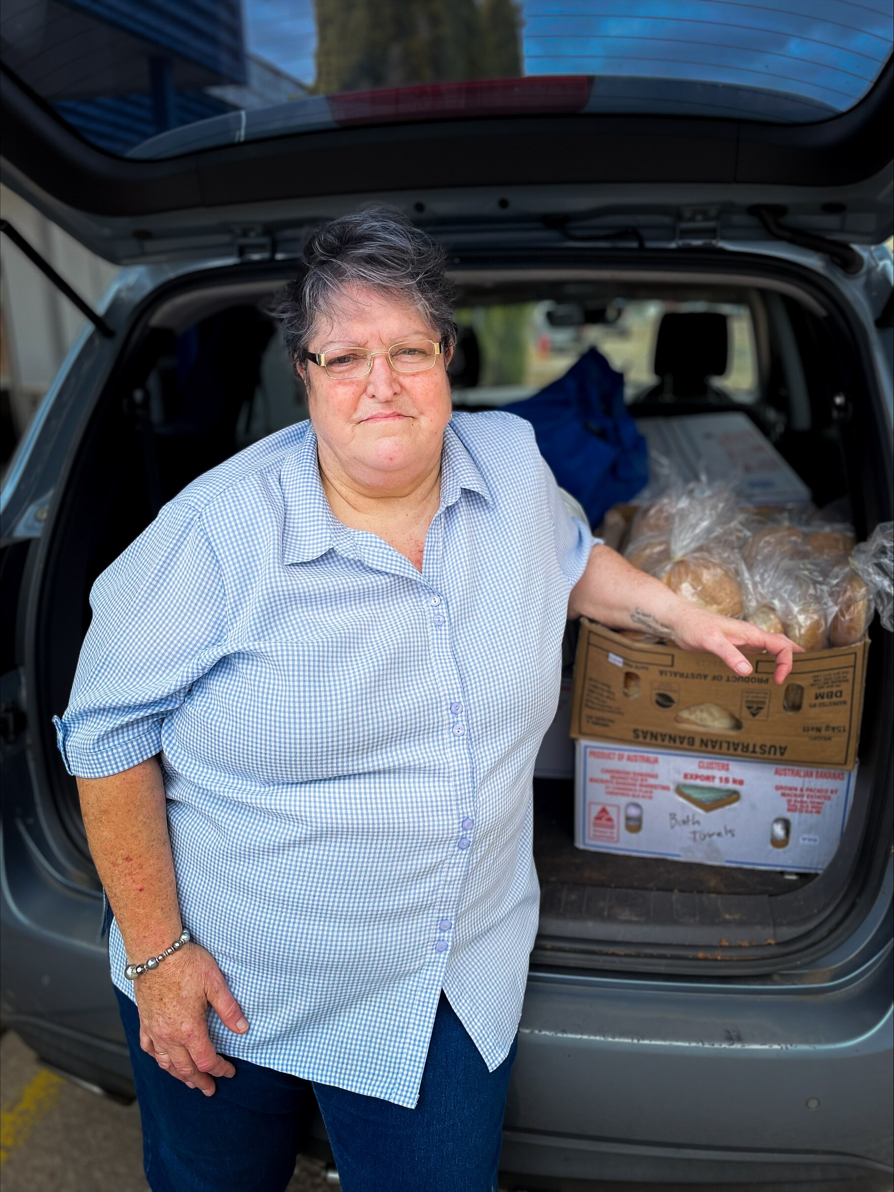 A woman stands in front of the open boot of her car, which contains a cardboard box of food