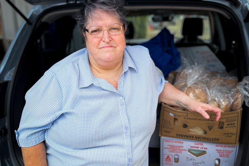 A woman stands in front of the open boot of her car, which contains a cardboard box of food