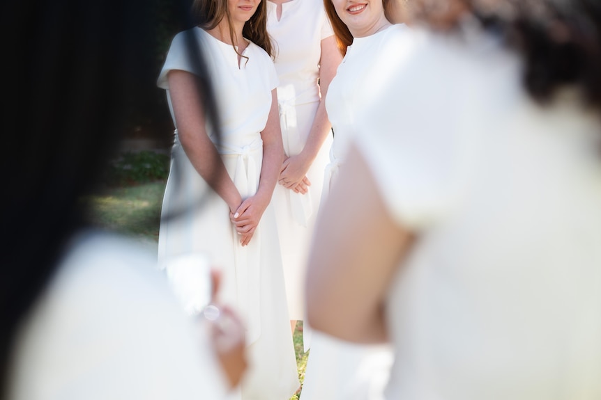 Five young women standing outside wearing matching white dresses.