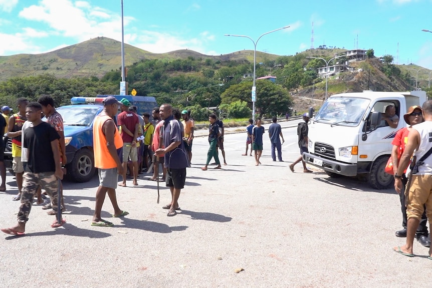 A group of people talk and walk around near a police car and a truck on a dirt road.