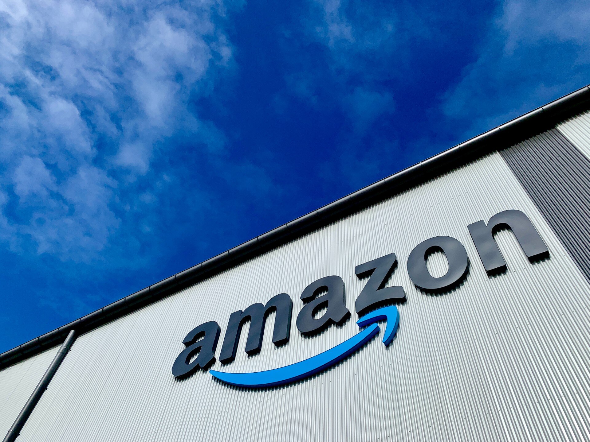 The Amazon logo is shown on the side of a building shot from below, with bright blue sky behind.