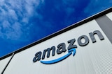 The Amazon logo is shown on the side of a building shot from below, with bright blue sky behind.