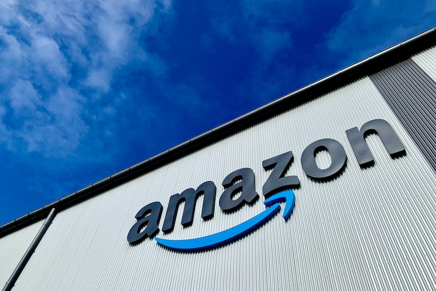 The Amazon logo is shown on the side of a building shot from below, with bright blue sky behind.