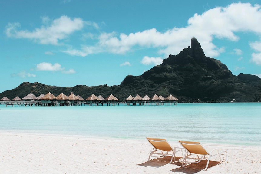 Two fold out chairs laid out on a sandy beach, overlooking clear waters and a mountain.