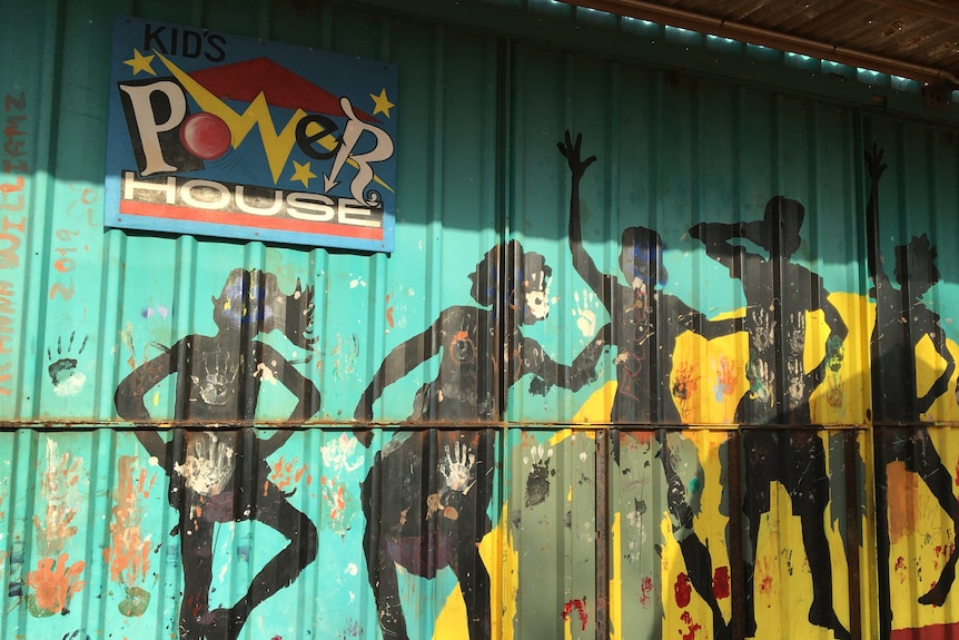 A mural painted on a tin shed depicting the silhouettes of five children playing and waving.