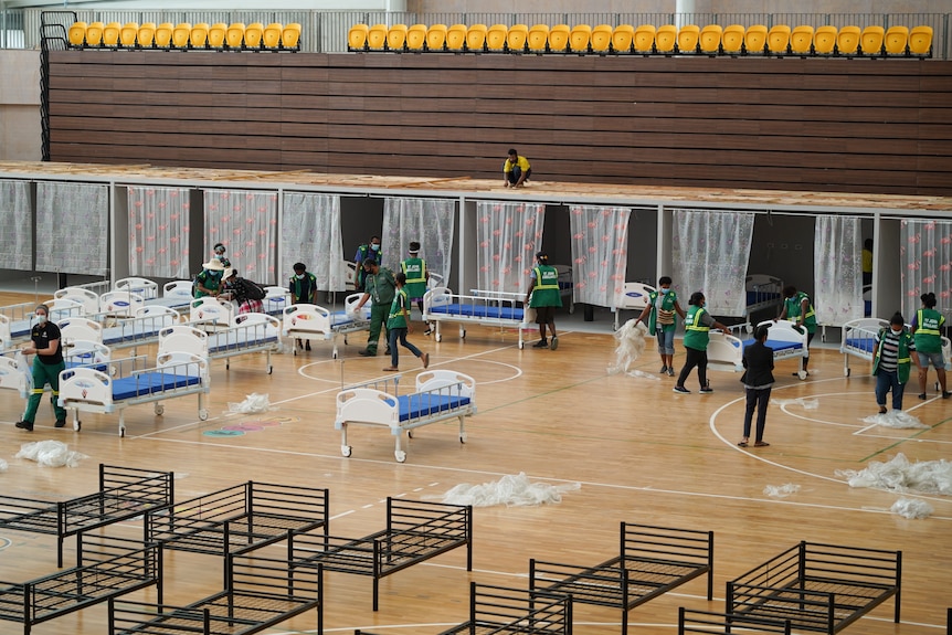 A group of people in green uniform and wearing masks walk around setting up beds in a room.