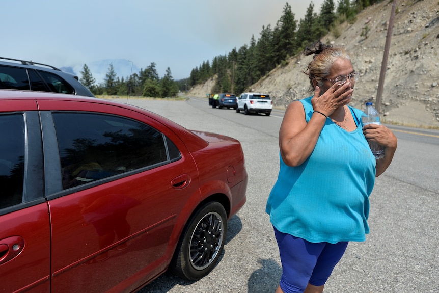 A woman stands next to a car on a rural road and wipes away tears.