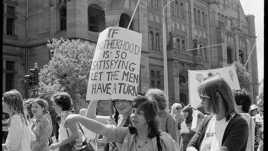 A black and white image of street protesters with a sign "if motherhood is so satisfying' let the men have a turn"