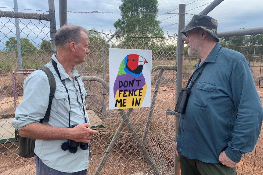 Two men with binoculars looking at a sign featuring a Gouldian finch, on a wire fence. 