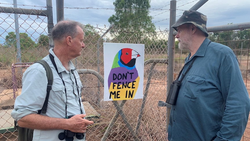 Two men with binoculars looking at a sign featuring a Gouldian finch, on a wire fence. 