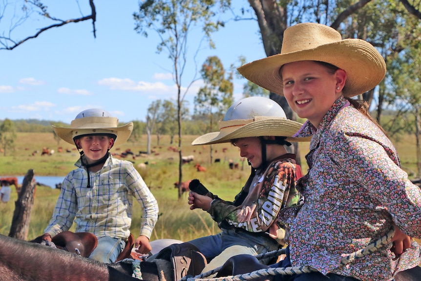 A girl and two boys sitting on their horses wearing cowboy hats and smiling