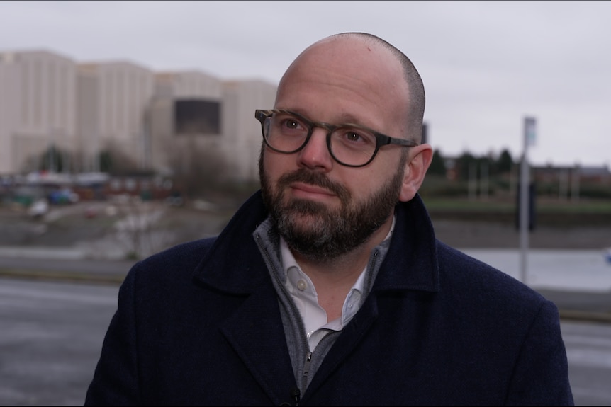 A man in glasses and a black coat looks towards the left. Behind him is a large industrial shipyard.