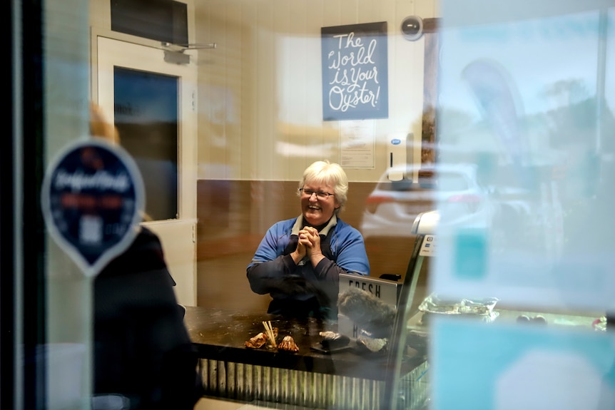 Woman with white hair wearing navy blue apron and blue shirt stands at shop counter smiling as she serves customers