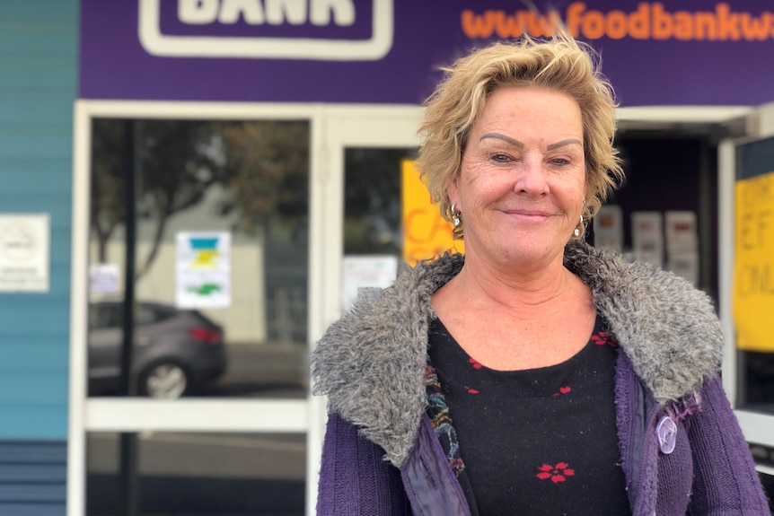 woman with short blonde hair and a grey jacket standing outside of purple bunbury foodbank building 
