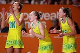 Three Australian netballers stand at courtside smiling and calling out support to their teammates during a game.