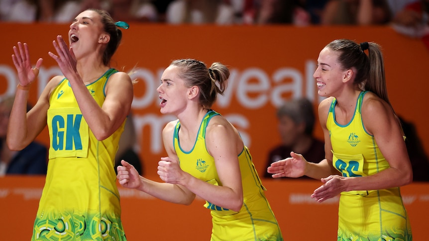Three Australian netballers stand at courtside smiling and calling out support to their teammates during a game.