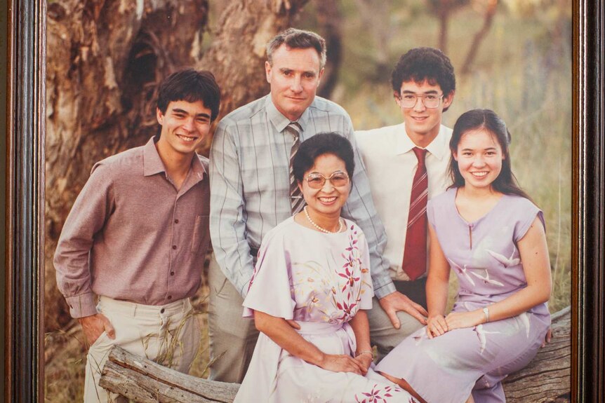 A 1980s family portrait of a man, woman and three teenagers or young adults in a photo frame on a wallpapered wall
