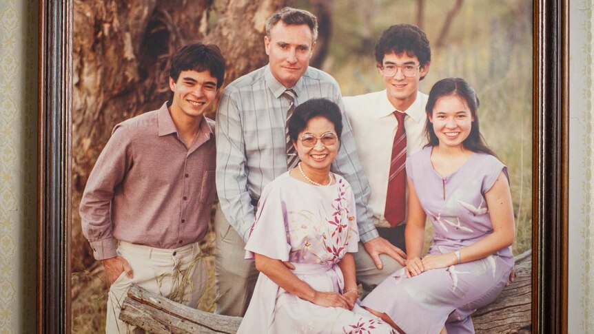 A 1980s family portrait of a man, woman and three teenagers or young adults in a photo frame on a wallpapered wall