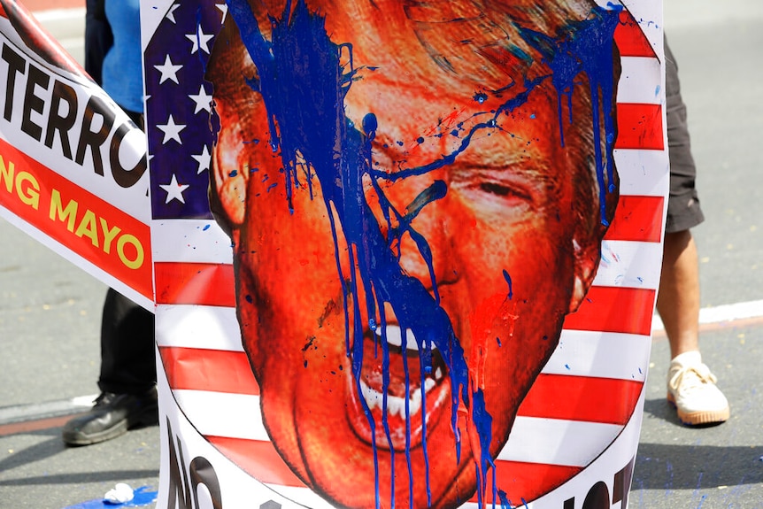 A persona holds placard of Donald Trump's face shouting is superimposed over a circular logo of the US flag splashed in blue.