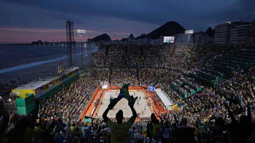 A view from the back of the stadium where a beach volleyball match is taking place.