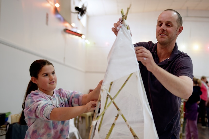 Man and young girl putting white paper on a bamboo lantern structure, both smiling. 