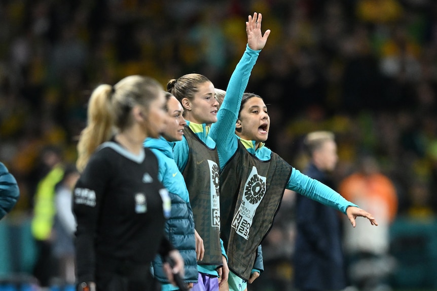 Sam Kerr shouts from a soccer pitch sideline, raising one hand and pointing with another. 