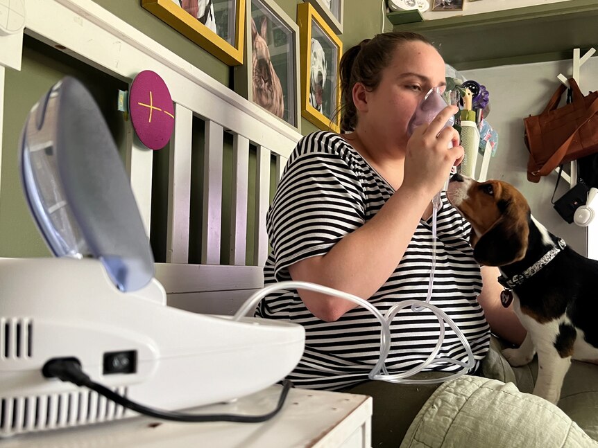 A brown-haired woman sits on her bed using asthma medication,