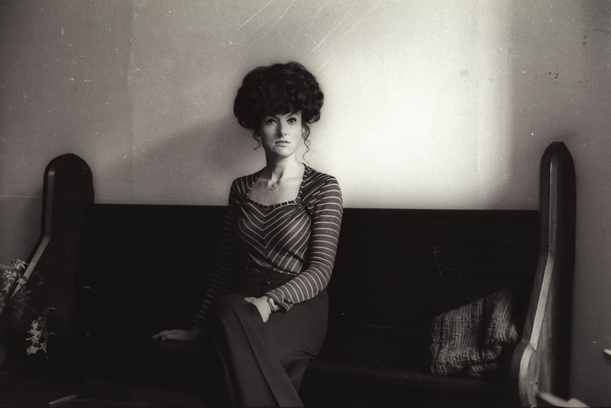 A black and white photo of a woman with a beehive hairdo seated and looking towards the camera.