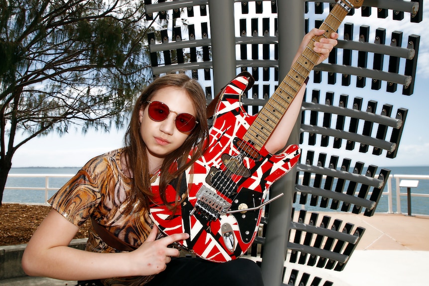 A girl with long hair, wearing circular sunglasses and holding an electric guitar pointing to the sky, in front of the beach