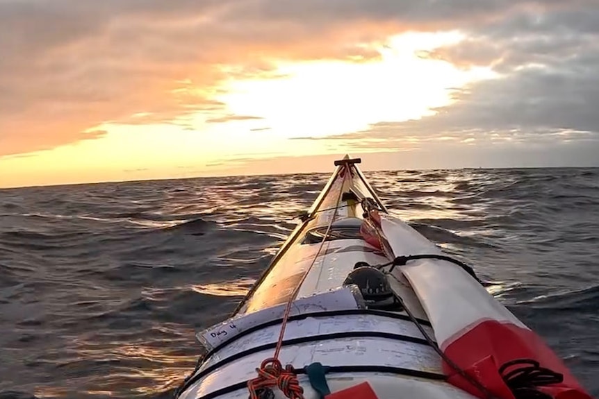 The hull of a white and red kayak in the water facing a sunset.