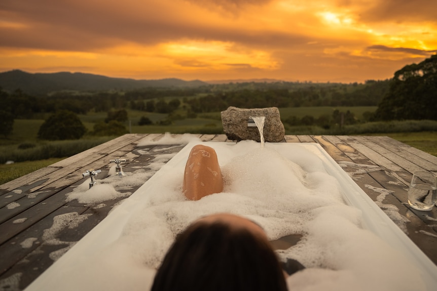 A woman lies in an outdoor bubble bath with a view out to a farm and sunset