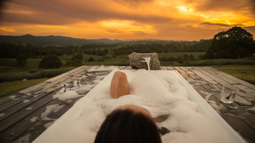 A woman lies in an outdoor bubble bath with a view out to a farm and sunset
