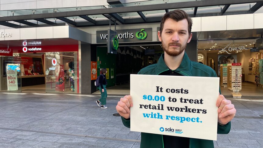 A man holding a sign in front of a supermarket, that says "it costs zero dollars to treat retail workers with respect".