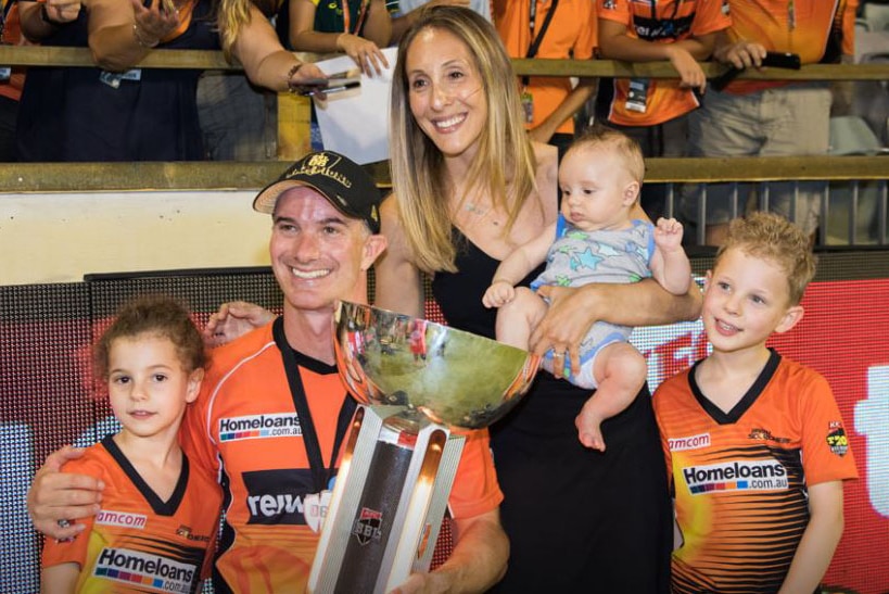 Cindy smiles while holding her baby, her husband kneels in front holding a trophy. Two other children flank either side.