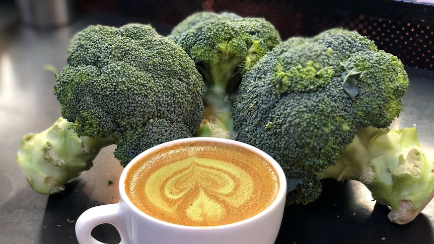 a small mug of flat white coffee in front of three heads of broccoli