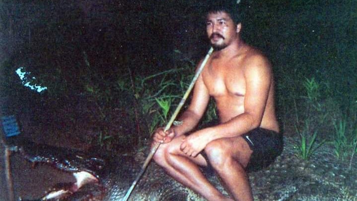 An Indigenous man sitting on a crocodile with a spear in his hand.
