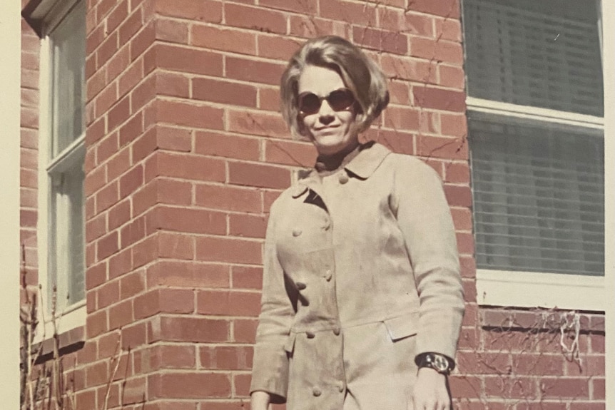 A woman with a fashionable 1960's bob haircut wearing sunglasses and a trench-style coat is smiling down at the camera