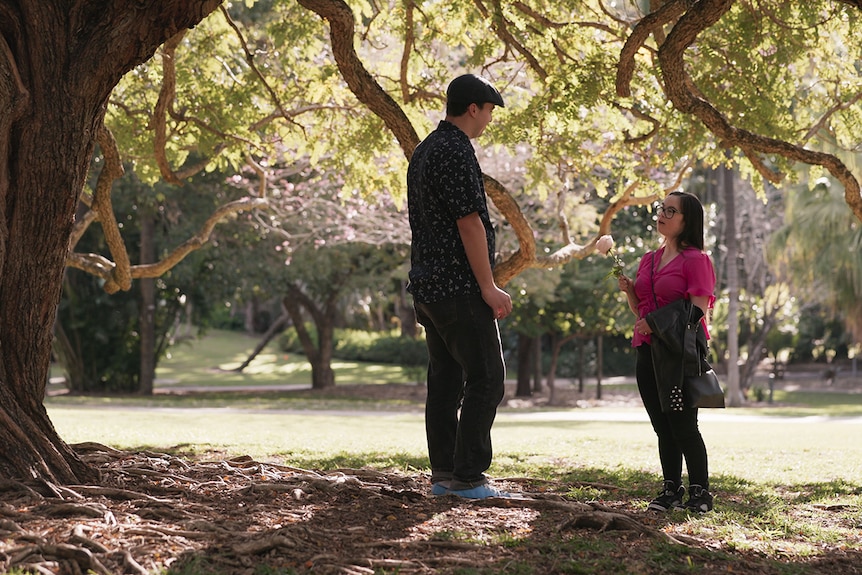 A man and woman stand under a big tree. The man is wearing dark colours and a hat, the woman is in a pink shirt and glasses.